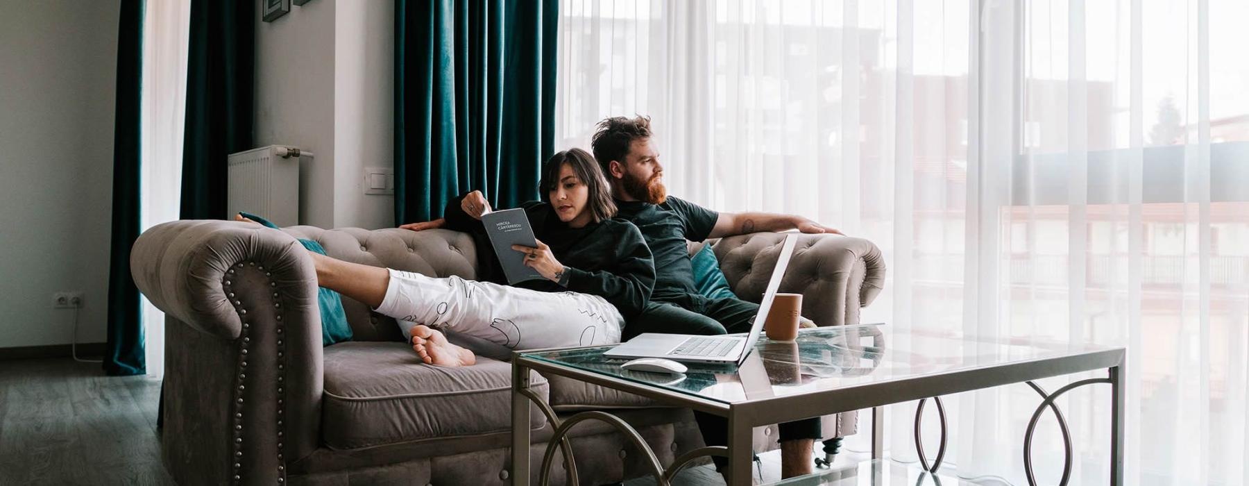 man sits on a couch and looks out the window while his wife lies against him reading a book